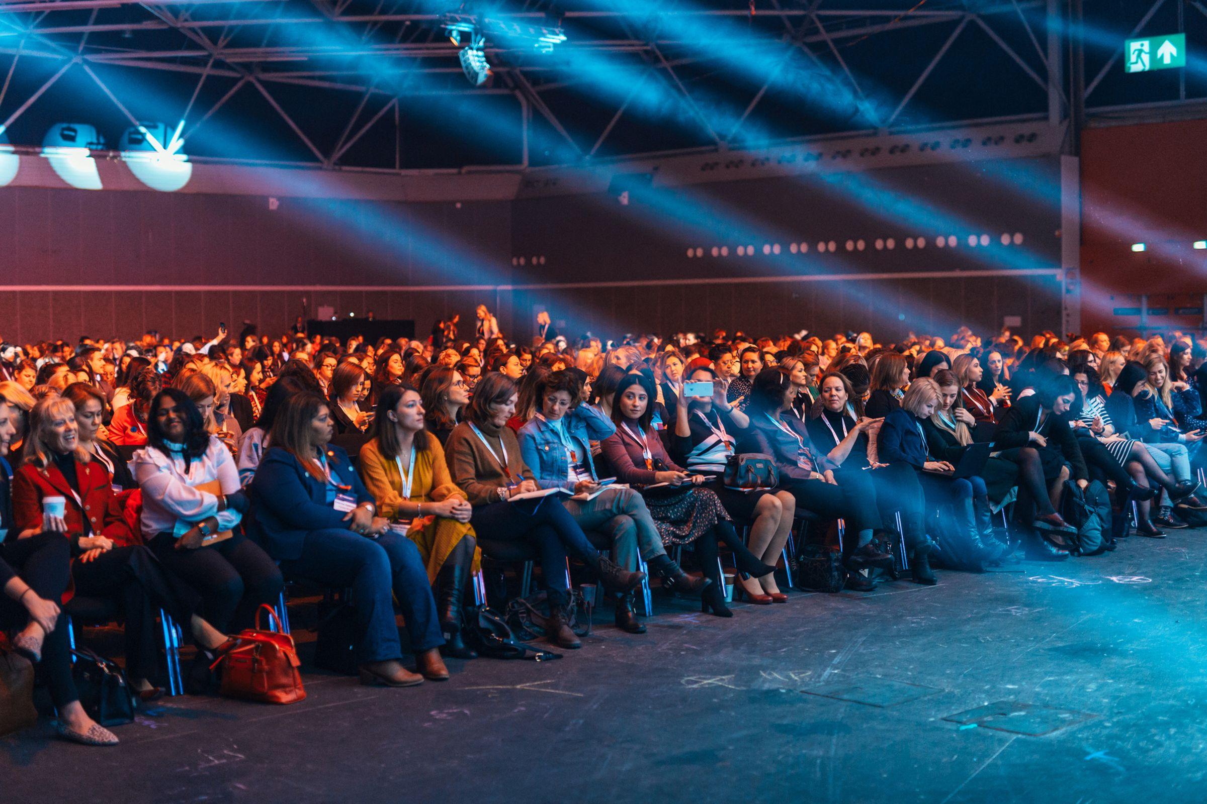A seated audience in a large room with light beams overhead.