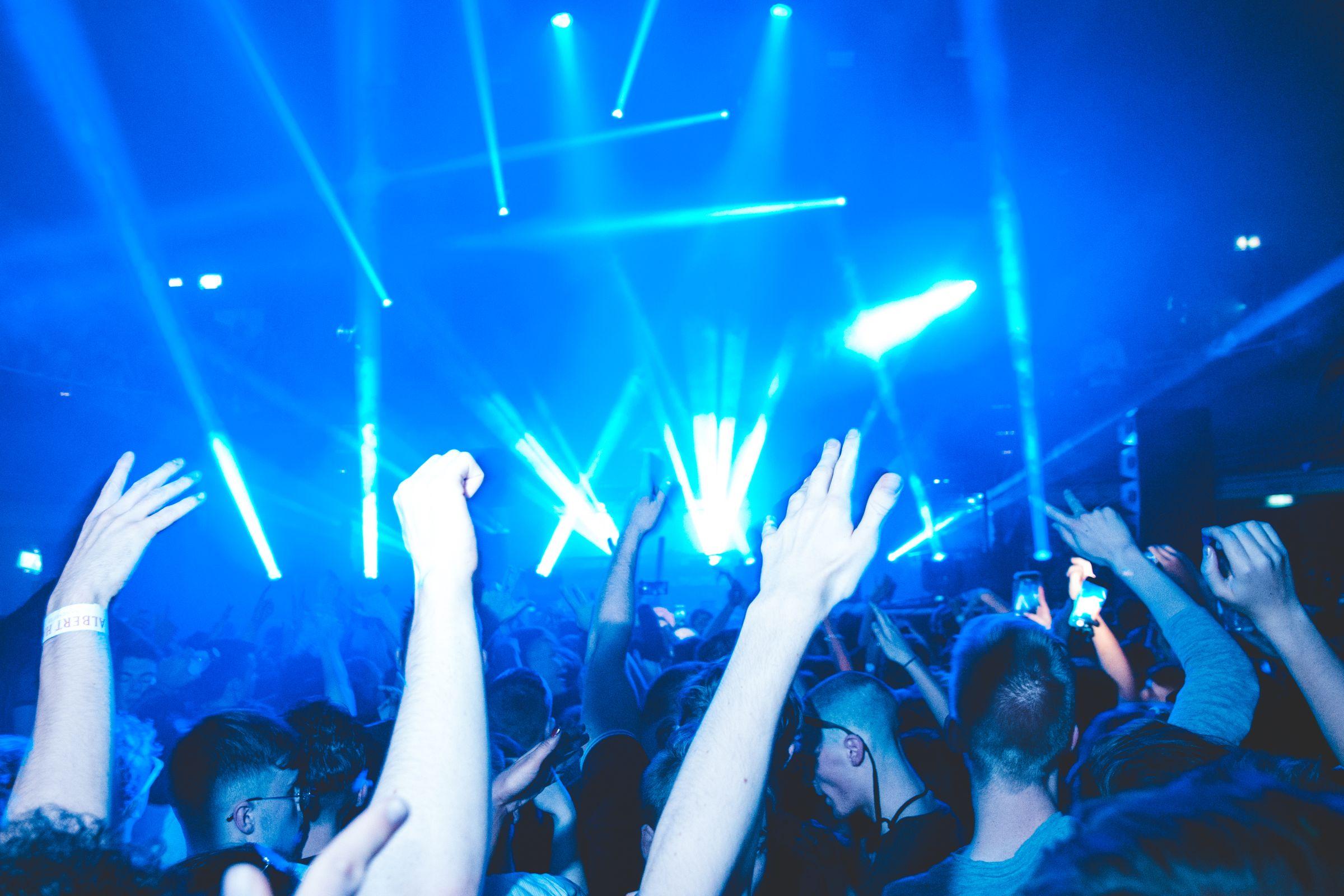 A club crowd with hands raised beneath sweeping light beams.