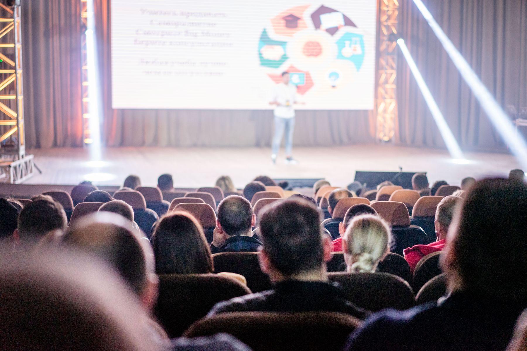 An audience watching a presentation under stage lighting.