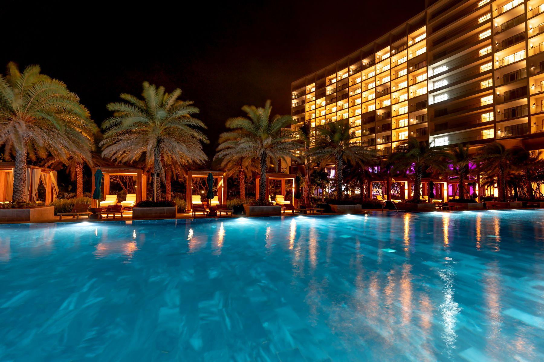 A resort pool lit at night, framed by palms and warm building light.